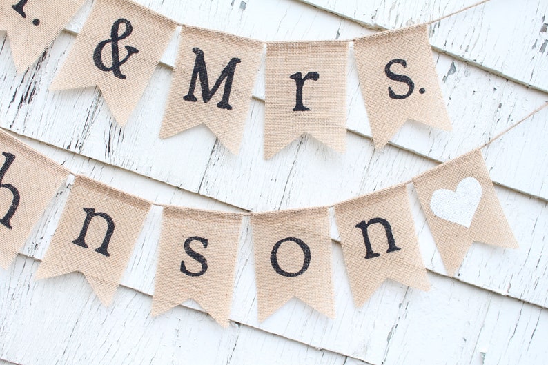 K&ouml;nnte beinhalten: Ein Banner aus Jute mit den Worten "Mr. & Mrs. Johnson" in schwarzer Schrift und einem wei&szlig;en Herz auf der letzten Flagge. Das Banner ist an einer wei&szlig;en Holzwand befestigt.