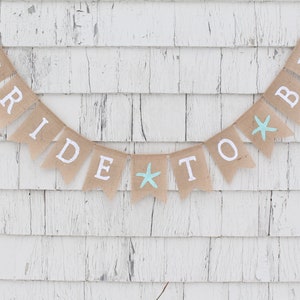 May include: A burlap banner with the words "Bride to Be" in white letters and two blue starfish accents. The banner is hanging on a white wooden wall.