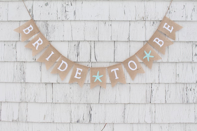 May include: A burlap banner with the words "Bride to Be" in white letters and two blue starfish accents. The banner is hanging on a white wooden wall.