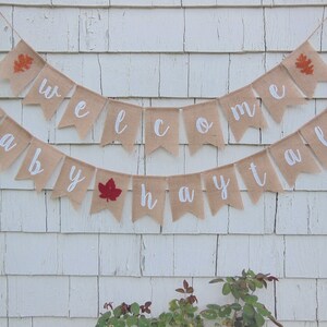 May include: A burlap banner with the words "Welcome Baby Fall" in white lettering. The banner is decorated with small orange and red felt leaves.