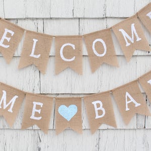 May include: A burlap banner with the words "Welcome Home Baby" in white lettering. A light blue heart is between the words "Home" and "Baby".