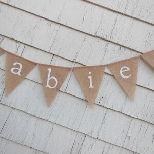 May include: A burlap banner with the word "babies" spelled out in white letters. The banner is hanging on a white wooden wall.