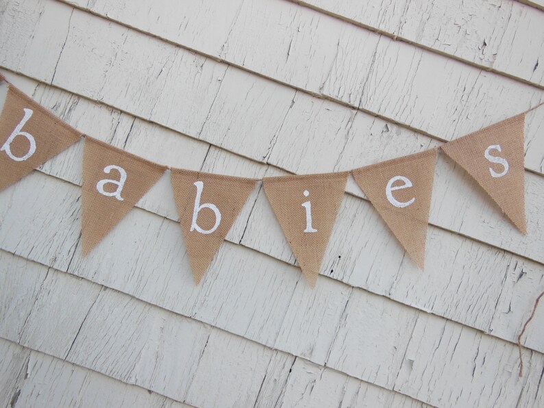 May include: A burlap banner with the word "babies" spelled out in white letters. The banner is hanging on a white wooden wall.