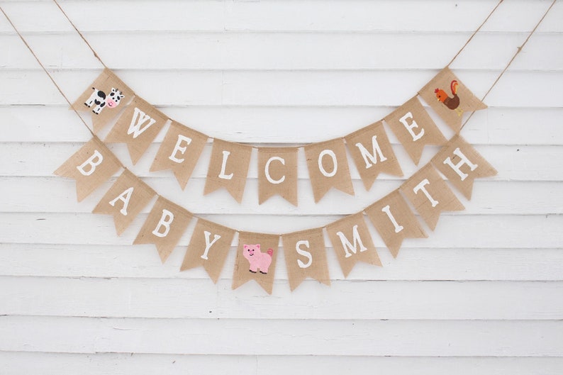 Pode incluir: Uma faixa de serapilheira com as palavras "WELCOME BABY SMITH" em letras mai&uacute;sculas brancas. A faixa &eacute; decorada com uma vaca, um porco e um galo. Pendurada por barbante contra um fundo de madeira branca.