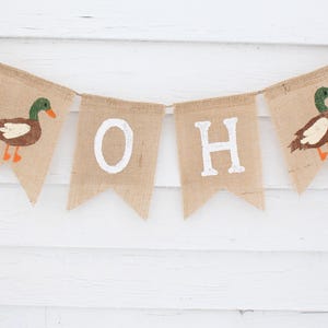May include: A burlap banner with the words "Oh Boy" in white paint. The banner is decorated with two brown and green ducks with orange beaks and feet.