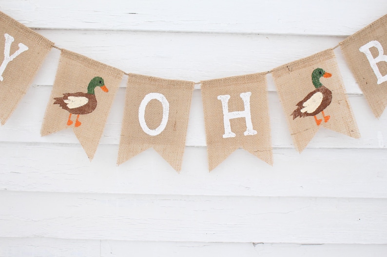 May include: A burlap banner with the words "Oh Boy" in white paint. The banner is decorated with two brown and green ducks with orange beaks and feet.