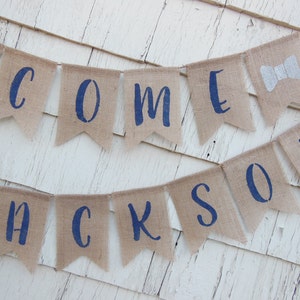 May include: Welcome banner with the name Jackson in navy blue letters on a burlap background. The banner is strung with twine and has a bow tie on the end.