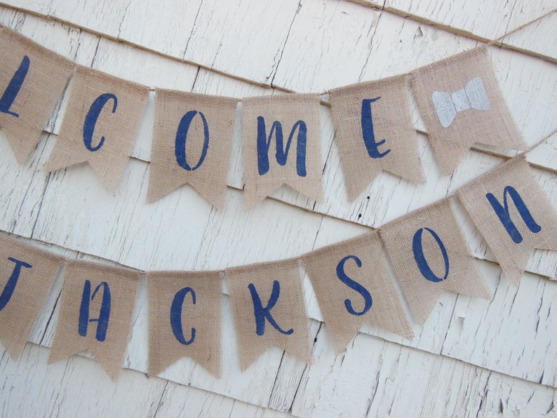 May include: Welcome banner with the name Jackson in navy blue letters on a burlap background. The banner is strung with twine and has a bow tie on the end.