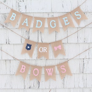 May include: A burlap banner with the words "BADGES OR BOWS" in light blue and pink lettering. The banner includes a police badge and a pink bow illustration. The banner is strung on twine against a white, weathered wood background.