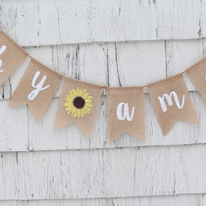 May include: A burlap banner with the words "Welcome" spelled out in white letters. A yellow sunflower is in the center of the banner.