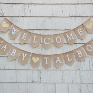 Puede incluir: Una pancarta de arpillera con las palabras "Welcome Baby Talbot" en letras blancas y corazones de purpurina dorada. La pancarta est&aacute; colgada en una pared de madera blanca.