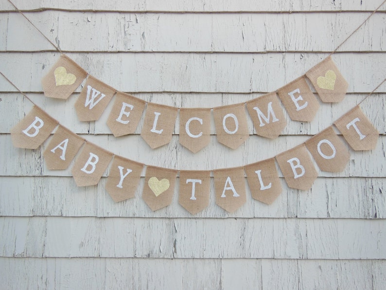 Puede incluir: Una pancarta de arpillera con las palabras "Welcome Baby Talbot" en letras blancas y corazones de purpurina dorada. La pancarta est&aacute; colgada en una pared de madera blanca.