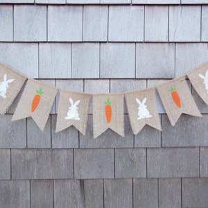 May include: A burlap banner with alternating images of white bunnies and orange carrots. The banner is strung up against a light brown wood background.