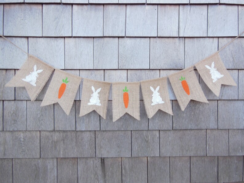 May include: A burlap banner with alternating images of white bunnies and orange carrots. The banner is strung up against a light brown wood background.
