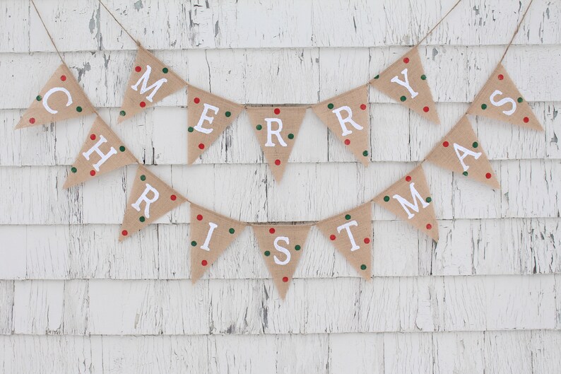 May include: A festive burlap banner spelling out "MERRY CHRISTMAS" in white capital letters. Each triangular flag is adorned with red and green polka dots. The banner is suspended on twine against a white, weathered wooden backdrop.