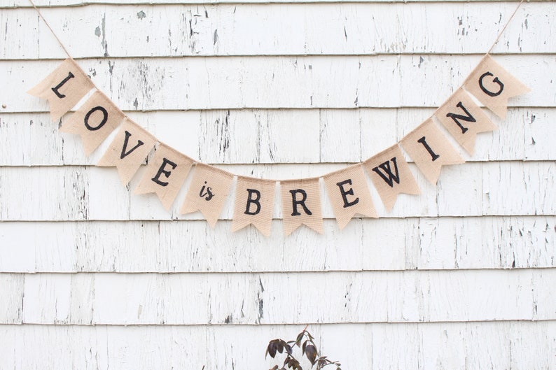 May include: A burlap banner with the text "LOVE is BREWING" in black lettering. The banner is strung up on a white wooden wall.
