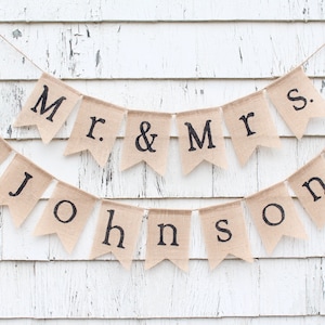 K&ouml;nnte beinhalten: Ein Banner aus Jute mit dem Text "Mr. & Mrs. Johnson" in schwarzen Buchstaben. Das Banner ist mit zwei wei&szlig;en Herzen verziert.