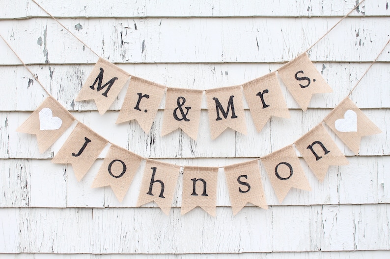 K&ouml;nnte beinhalten: Ein Banner aus Jute mit dem Text "Mr. & Mrs. Johnson" in schwarzen Buchstaben. Das Banner ist mit zwei wei&szlig;en Herzen verziert.