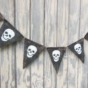 May include: A black burlap banner with white skull designs. The pennant-shaped flags are strung together, creating a festive decoration. The skulls are a classic Halloween or pirate theme.