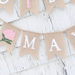 Puede incluir: Una pancarta de arpillera con la palabra "MAY" en letras blancas y una flor rosa con hojas verdes en una de las banderas.
