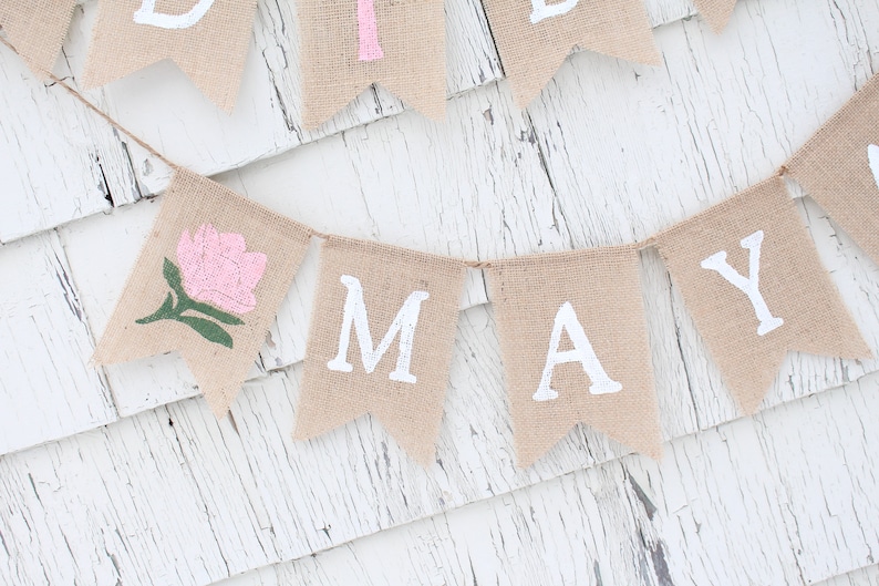 Puede incluir: Una pancarta de arpillera con la palabra "MAY" en letras blancas y una flor rosa con hojas verdes en una de las banderas.
