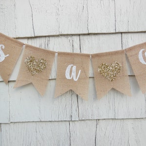 May include: A burlap banner with the words "It's a girl" in white lettering. The banner has gold glitter hearts between each letter.