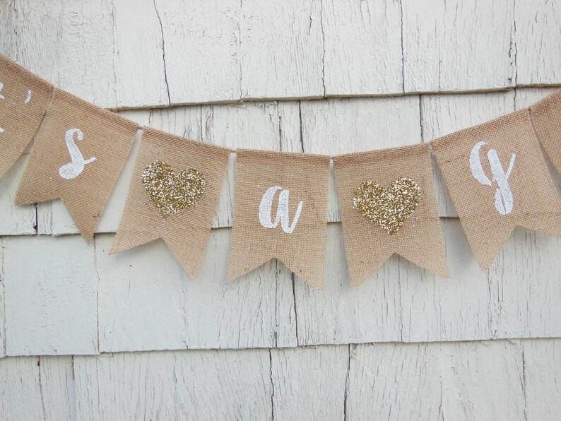 May include: A burlap banner with the words "It's a girl" in white lettering. The banner has gold glitter hearts between each letter.