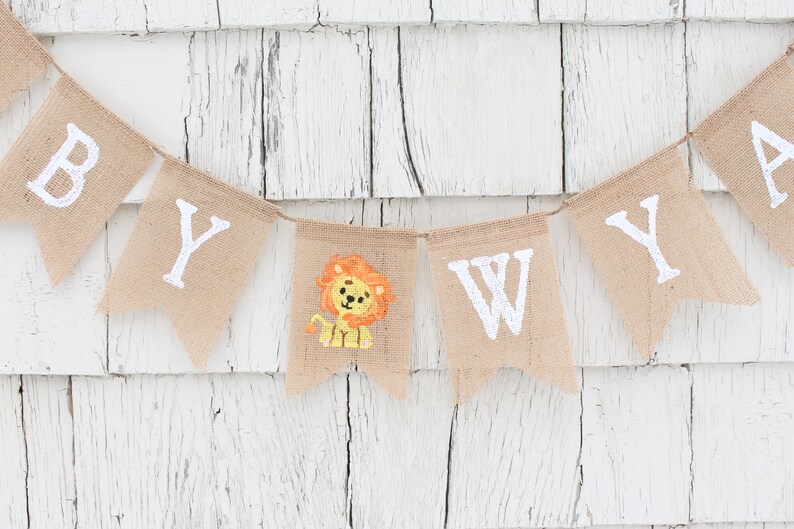 Pode incluir: Uma faixa de juta com as palavras "Baby Wyatt" pintadas de branco. Uma pequena ilustra&ccedil;&atilde;o de le&atilde;o fica entre as palavras "Baby" e "Wyatt".