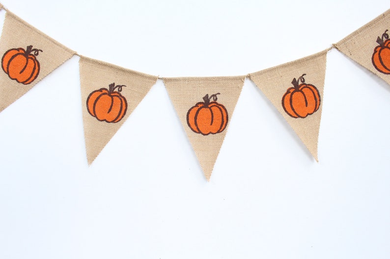 May include: A burlap banner with triangular pennants, each displaying an orange pumpkin design with a brown stem. This banner is suitable for autumn or fall decorations. The pumpkins are centered on the pennants.