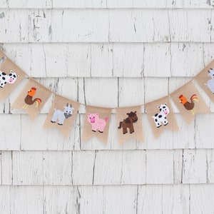 May include: A burlap banner with farm animal illustrations. The banner features a cow, pig, horse, goat, rooster, and a brown and white cow. The banner is strung on a white wall.