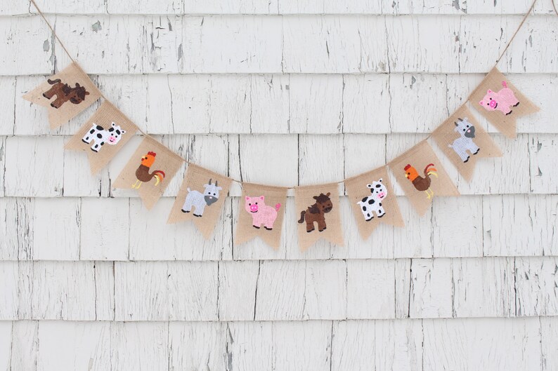 May include: A burlap banner with farm animal illustrations. The banner features a cow, pig, horse, goat, rooster, and a brown and white cow. The banner is strung on a white wall.