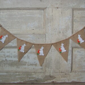 May include: A burlap banner with white bunny silhouettes holding carrots. The banner is strung together with white string and is perfect for an Easter party or celebration.