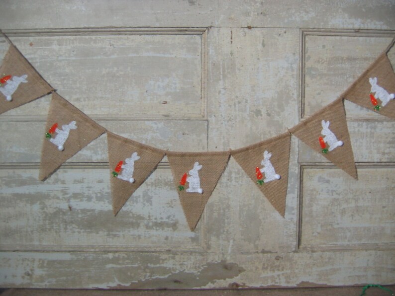 May include: A burlap banner with white bunny silhouettes holding carrots. The banner is strung together with white string and is perfect for an Easter party or celebration.