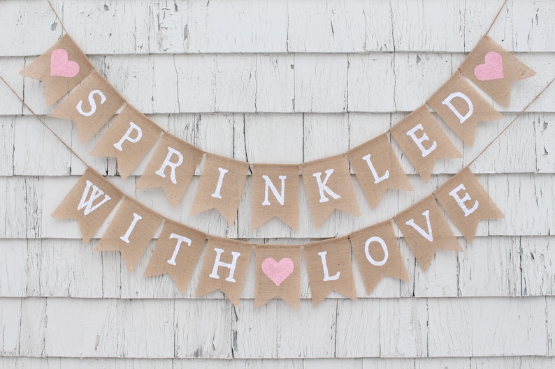 May include: A burlap banner with pink hearts and the text "Sprinkled with Love" in white. The banner is hanging on a white wooden wall.