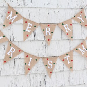May include: A festive burlap banner spelling "MERRY CHRISTMAS" in white capital letters. Each triangular flag is adorned with red and green dots. The banner is suspended on twine against a white, weathered wooden backdrop.