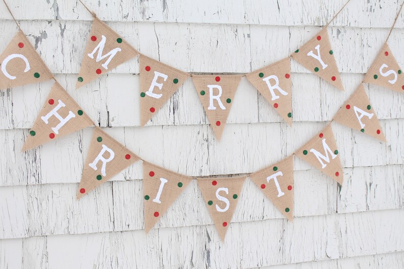 May include: A festive burlap banner spelling "MERRY CHRISTMAS" in white capital letters. Each triangular flag is adorned with red and green dots. The banner is suspended on twine against a white, weathered wooden backdrop.