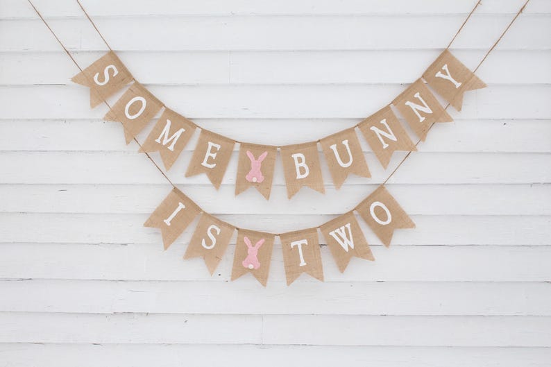 May include: A burlap banner with the text "Some Bunny Is Two" in white letters. The banner is decorated with pink bunny silhouettes.