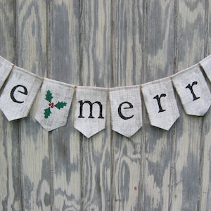 May include: A burlap banner with the words "be merry" in black letters. The letter "e" has a sprig of holly with red berries and green leaves. The banner is strung on a natural twine and hangs against a weathered wood background.