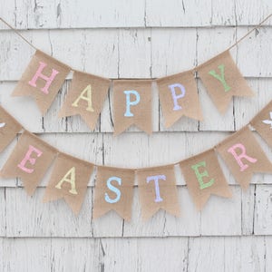 May include: A burlap banner with the words "Happy Easter" in pastel colors. The banner is strung up on a white wooden wall.