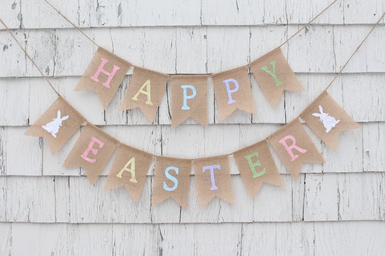 May include: A burlap banner with the words "Happy Easter" in pastel colors. The banner is strung up on a white wooden wall.