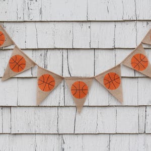 Puede incluir: Una pancarta de arpillera con balones de baloncesto naranjas impresos en cada bandera triangular. La pancarta est&aacute; colgada en una pared de madera blanca.