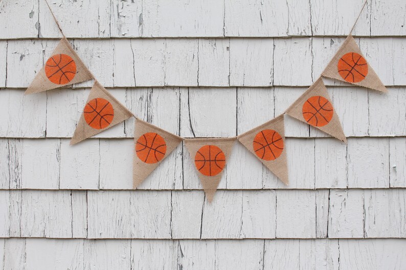 Puede incluir: Una pancarta de arpillera con balones de baloncesto naranjas impresos en cada bandera triangular. La pancarta est&aacute; colgada en una pared de madera blanca.