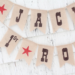Pode incluir: Bandeirola de juta com o nome "JACK ROD" e o n&uacute;mero "1st" impressos em castanho escuro. Estrelas vermelhas tamb&eacute;m est&atilde;o inclu&iacute;das na bandeirola. A bandeirola est&aacute; pendurada em uma corda fina e est&aacute; sobre um fundo de madeira branca.