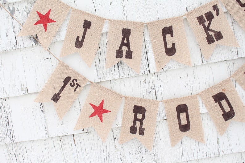 Pode incluir: Bandeirola de juta com o nome "JACK ROD" e o n&uacute;mero "1st" impressos em castanho escuro. Estrelas vermelhas tamb&eacute;m est&atilde;o inclu&iacute;das na bandeirola. A bandeirola est&aacute; pendurada em uma corda fina e est&aacute; sobre um fundo de madeira branca.