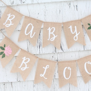 May include: A burlap banner with the words "Baby Bloom" in white paint. The banner is decorated with pink flowers and green leaves.
