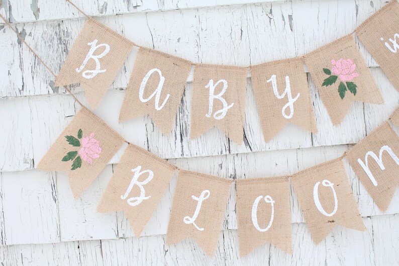May include: A burlap banner with the words "Baby Bloom" in white paint. The banner is decorated with pink flowers and green leaves.