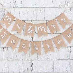 K&ouml;nnte beinhalten: Ein Banner aus Jute mit den Worten "Mr. & Mrs. Jackson" in wei&szlig;en Buchstaben und zwei rosa Herzen. Das Banner ist an einer wei&szlig;en Holzwand befestigt.