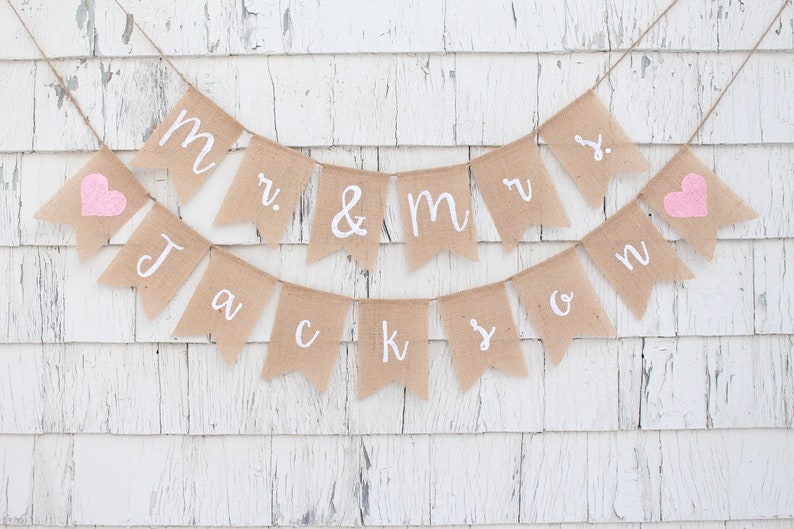 K&ouml;nnte beinhalten: Ein Banner aus Jute mit den Worten "Mr. & Mrs. Jackson" in wei&szlig;en Buchstaben und zwei rosa Herzen. Das Banner ist an einer wei&szlig;en Holzwand befestigt.
