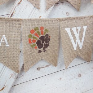 May include: A burlap banner with white painted letters and a turkey graphic. The banner spells out "YA" and "W". The turkey graphic is in shades of brown, orange, and green. The banner is hanging against a white wooden background.