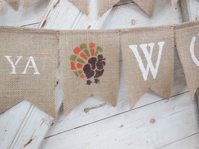 May include: A burlap banner with white painted letters and a turkey graphic. The banner spells out "YA" and "W". The turkey graphic is in shades of brown, orange, and green. The banner is hanging against a white wooden background.